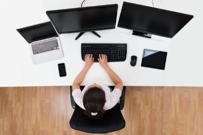 Top down view: woman sitting in front of two computer monitors, and one laptop to her left. A tablet sits to her right. Her hands rest on the keyboard.