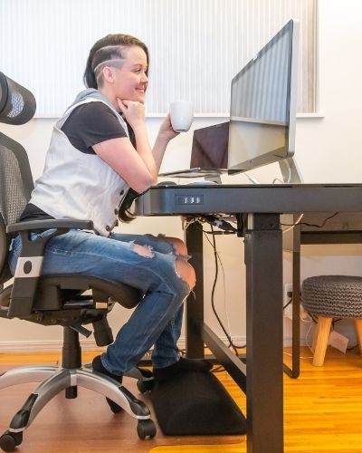 Woman sitting in desk chair, at desk, holding coffee, looking at computer monitor